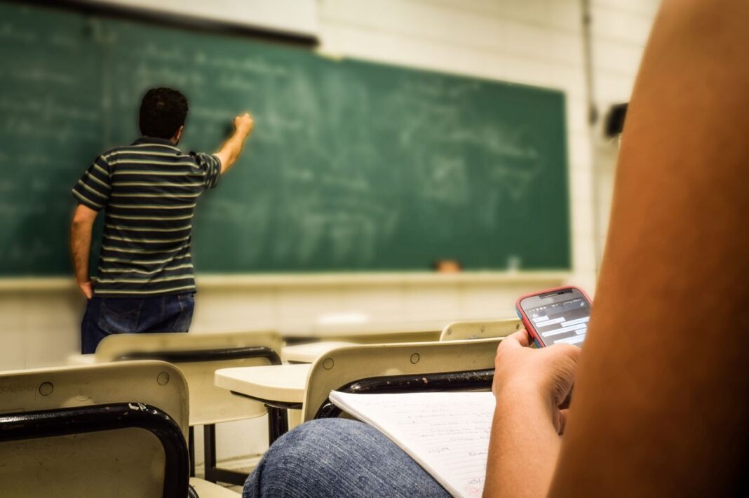Profesor dando clase y escribiendo en la pizarra mientras un alumno usa su teléfono móvil