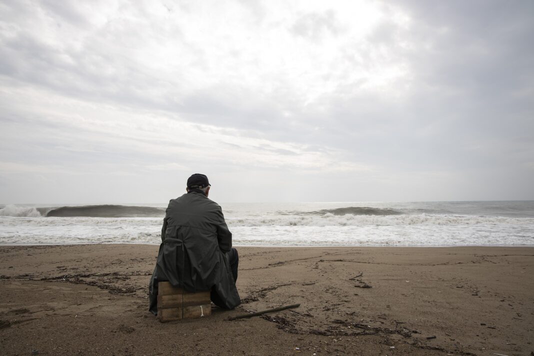 Hombre con depresión sentado en la orilla de la playa observando melancólico la inmensidad del mar